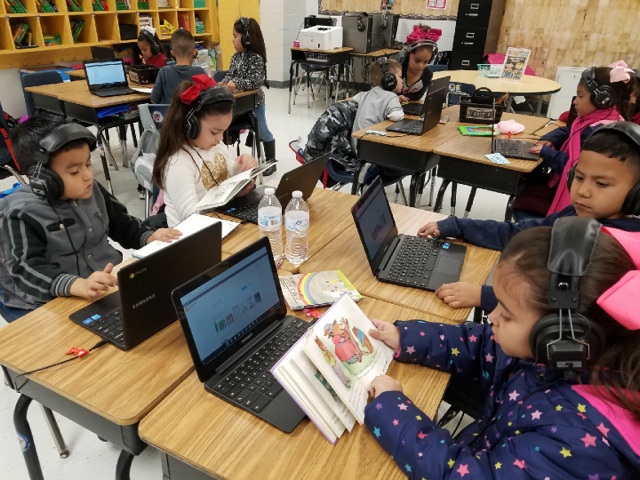 Students using technology and following along with a book Students using technology and following along with a book